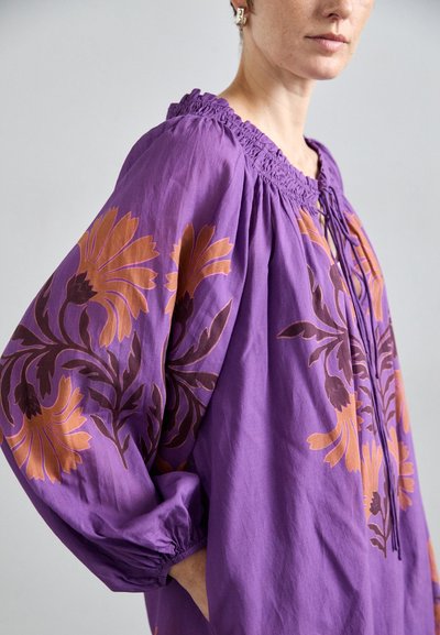 Paarse katoenen blouse met een gesmokte halslijn, lange mouwen en bloemige bruine en oranje patronen op de mouwen. Textuurstof.