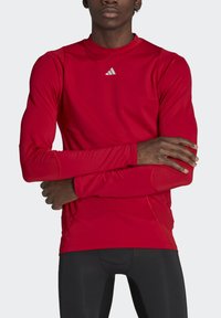 Rotes Sportshirt mit langen Ärmeln aus strukturiertem Stoff, mit Rundhalsausschnitt und kleinem silbernen Logo auf der Brust.