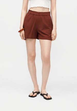 Kvinde iført højtaljede mørkerøde shorts, hvid croppet top, brun armbånd og sorte flade sandaler mod en ensfarvet baggrund.