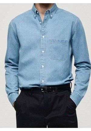 Homme portant une chemise en jean bleu clair à boutons avec une poche poitrine rentrée dans un pantalon noir avec ceinture noire, mains dans les poches.