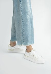 Sneaker bianco con dettagli dorati e lati texture, abbinati a denim azzurro chiaro sfilacciato, con una striscia di paillettes lucida lungo la gamba dei pantaloni.
