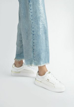 Sneaker bianco con dettagli dorati e lati texture, abbinati a denim azzurro chiaro sfilacciato, con una striscia di paillettes lucida lungo la gamba dei pantaloni.