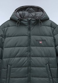Mørkegrøn pufferjakke med kviltet design, der har lynlås og to sidelommer. Inkluderer hætte med justerbare snore.