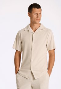 Camicia a maniche corte beige con bottoni in tessuto morbido e peloso, dotata di colletto. Presenta due tasche frontali e una vestibilità casual, abbinata a pantaloni coordinati.