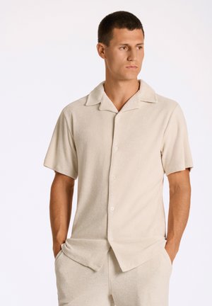 Camisa de botones de manga corta en color beige, fabricada en un tejido suave y aterciopelado, con cuello. Presenta dos bolsillos frontales y un corte relajado, combinada con pantalones a juego.