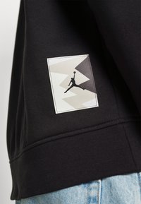 Felpa nera con una patch logo bianca e grigia raffigurante una figura che salta sul lato sinistro, su uno sfondo di tessuto liscio.