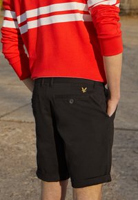 Sorte shorts med ombukket kant, forlomme og en baglomme med knap, der har et lille guldørnemblem. Stoffet ser glat ud.