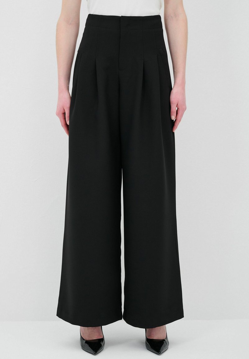 Pantalons larges noirs à taille haute, avec des plis à l'avant et des poches sur les côtés. Le tissu est lisse et fluide, complété par des talons aiguilles noirs.