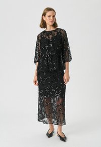 Robe noire en sequins avec superposition en transparence, dotée de larges manches 3/4 et d'un design superposé. Longue jusqu'au sol avec une silhouette fluide.