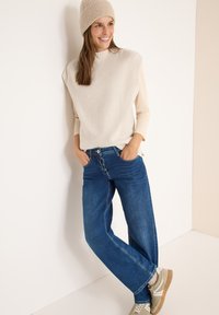 Beige gerippter Strickpullover, weite blaue Jeans, hellgraue Sneakers und eine passende Strickmütze. Schlichtes Design mit lockerer Passform.