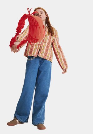 Ung kvinde i blomstrede stribede skjorte og vidde jeans, der svinger en rød skuldertaske mod kameraet mod hvid baggrund.