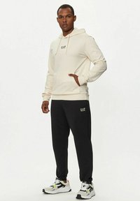 Felpa beige con tasca a marsupio e logo, abbinata a pantaloni della tuta neri con logo, indossata con sneakers bianche decorate con dettagli in neon.