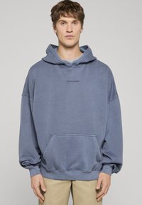 Nadrozmerná modrá mikina s kapucňou, predným vreckom, rebrovanými manžetami a vyšitým logom na hrudi. Vyrobená z mäkkej, textúrovanej látky.