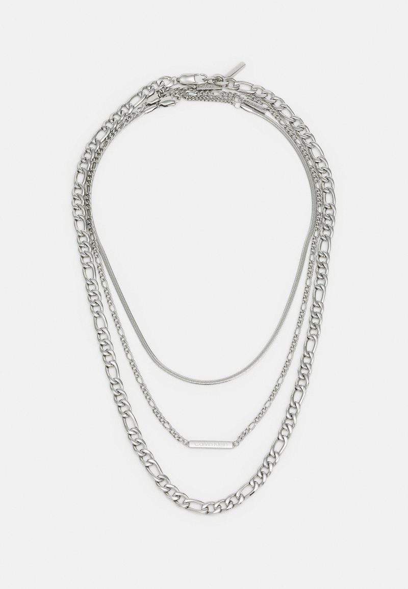 Collier en chaîne argenté à plusieurs couches présentant des épaisseurs et des designs variés, y compris une chaîne à maillons plats et une chaîne en chevrons lisse.