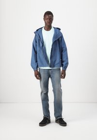 Jeansjakke med hette, glidelåslukking og snøring. Lyseblå t-skjorte under. Bært med lyseblå jeans og svarte joggesko.