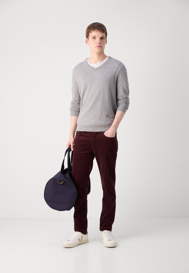 Grauer V-Ausschnitt-Pullover, burgunderfarbene Cordhosen, weiße Sneaker mit beige Akzenten und eine runde marineblaue Tasche mit Griffen. Glatte Textur, lässiges Design.