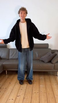 Crna, pahuljasta jakna s kapuljačom preko bijele majice, u kombinaciji s širokim plavim trapericama i crnim cipelama. Drveni pod i siva sofa u pozadini.