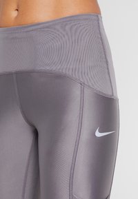 Leggings deportivos de talle alto en gris, fabricados con un material suave y elástico. Presentan un logo de Nike plateado y una cinturilla texturizada.