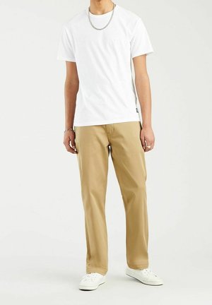T-shirt en coton blanc associé à un chino beige. Le modèle porte des baskets blanches et une chaîne en argent. Design épuré, coupe décontractée et longueur moyenne.