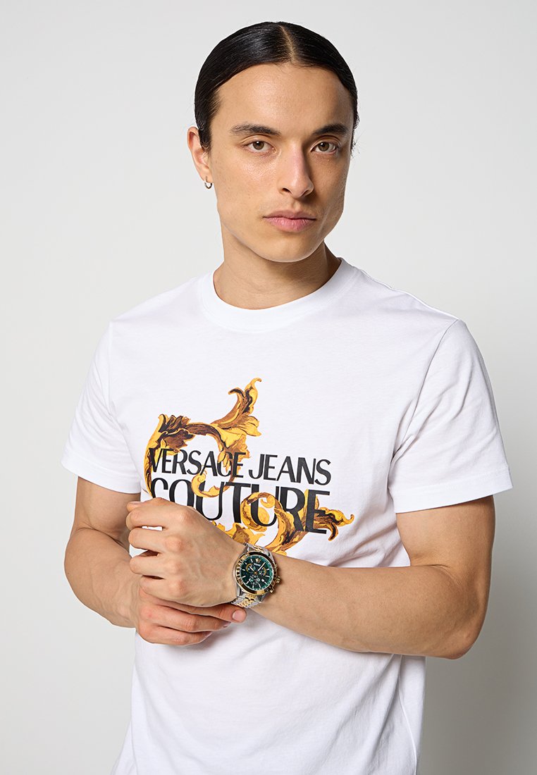 T-shirt bianco con il testo "VERSACE JEANS COUTURE", abbinato a motivi barocchi dorati. Il modello indossa un orologio verde e argento.