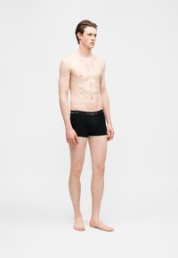 Νεαρός άνδρας όρθιος, ξυπόλητος, φοράει μαύρα μποξεράκια με λογότυπο στη μέση, στραμμένος ελαφρώς προς τα δεξιά, σε λευκό φόντο.