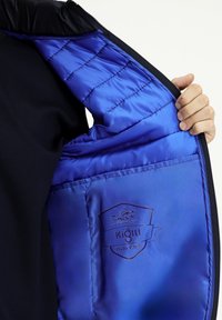 Czarna kurtka z niebieskim satynowym wnętrzem, z pikowanym szyciem i naszywką z logo "Kigili Polo Club" po lewej stronie.