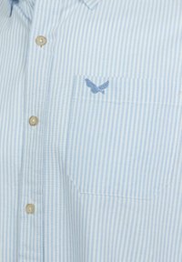 Camisa a rayas azul claro con cierre de botones en el frente, que presenta un pequeño detalle bordado de ala en el bolsillo del pecho y botones de color marrón claro.