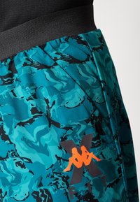 Teal un melnā krāsā rakstotas sporta šorti ar plašu melnu jostu; apakšējā labajā pusē ir oranžs logotipa detalizējums.