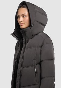 Sieviete valkā tumši pelēku ziemas jaku ar rāvējslēdzēju un lielu kapuci, skatās pa kreisi pret vienkāršu fonu.