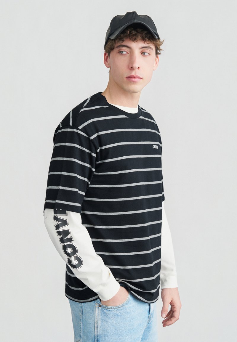 Νέος άνδρας που φοράει μαύρο ριγέ πουκάμισο με κοντά μανίκια πάνω από λευκό πουκάμισο με μακριά μανίκια και το κείμενο "CONV." στο μανίκι, ανοιχτόχρωμο τζιν και μαύρο καπέλο.