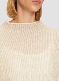 Helles beiges Strickpullover mit hohem, geripptem Kragen und subtiler Textur. Der Stoff wirkt weich und hat am Ausschnitt eine leicht durchscheinende Qualität.