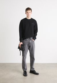 Crna dukserica s logotipom, sivi traperice, crni remen i crne tenisice. Drži malu crnu torbu. Jednostavna pozadina.