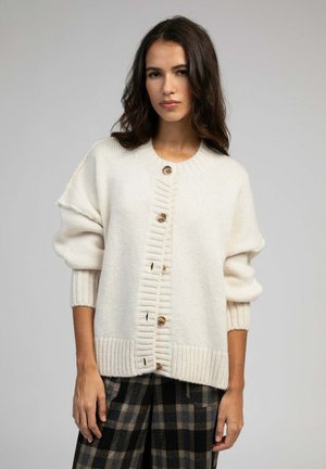 Cardigan tricoté crème avec des poignets et un ourlet côtelés, doté d'une fermeture à boutons et de détails texturés. Porté par-dessus un pantalon à carreaux.