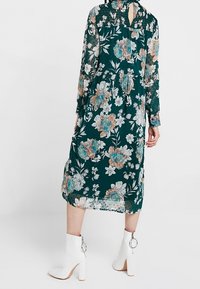Długa, kwiecista sukienka w ciemnozielonym kolorze z brzoskwiniowymi i niebieskimi kwiatami. Przezroczysty materiał, falbaniasty dekolt i detal marszczenia z tyłu.