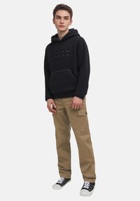 Hoodie en polaire noire avec une poche kangourou et un logo en relief, associé à un pantalon cargo beige et des baskets noires à semelles blanches.