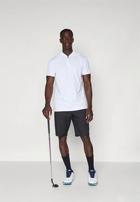 Biela polokošeľa s krátkym rukávom a zipsovým golierom v kombinácii s čiernymi šortkami, čiernymi ponožkami a bielymi golfovými topánkami, držiaca golfovú palicu.