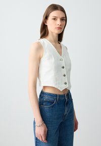 Gilet in denim bianco a vita corta con scollo a V, caratterizzato da cinque bottoni argentati e dettagli a strappo sul bordo, abbinato a jeans blu a vita alta.