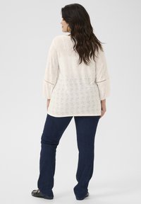 Crèmekleurige blouse met gaatjespatroon, uitlopende driekwartmouwen en een losse pasvorm, gecombineerd met donkerblauwe uitlopende spijkerbroeken en zwarte schoenen.