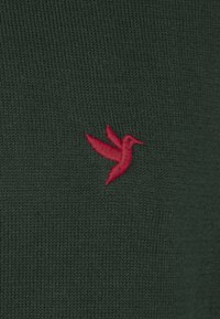 Donkergroen gebreide stof met een klein rood borduursel van een kolibrie aan de linkerkant. De textuur oogt glad en egaal.