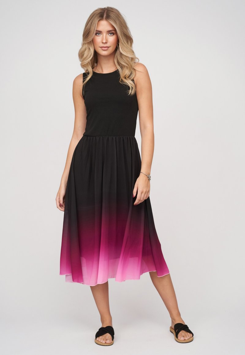 Femme aux longs cheveux blonds portant une robe noire sans manches avec une jupe dégradée rose et des sandales noires, debout devant un fond uni.