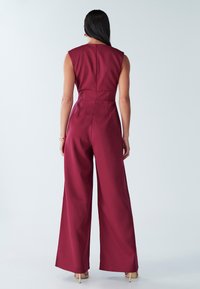 Mono color burdeos con diseño sin mangas, pantalones de pierna ancha y cintura ajustada. Tejido suave, cremallera visible en la espalda y detalles mínimos de costura.