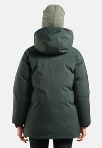 Doudoune vert foncé avec capuche, dotée d'une surface lisse, de poignets élastiques et d'une coupe mi-longue. Portée par-dessus un pantalon noir.