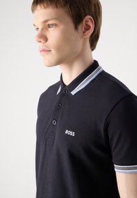 Crna polo majica s klasičnim krojem. Ima ovratnik s plavim prugastim obrubom i istaknutim bijelim "BOSS" logom na prsima.