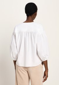 Frau mit geflochtenem Haar, das zu einem Dutt gebunden ist, trägt eine lockere weiße Bluse mit Puffärmeln und beigen Hosen, steht mit dem Rücken zu einem schlichten Hintergrund.