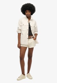 Chaqueta abotonada color beige claro con dos bolsillos en el pecho, camiseta negra y pantalones cortos blancos con dobladillo deshilachado, combinados con zapatillas beige.