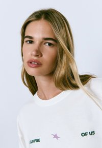 Mujer joven con cabello largo y rubio, vistiendo una camisa blanca con una estrella morada y el texto verde "UN VISTAZO A NOSOTROS" sobre un fondo claro.