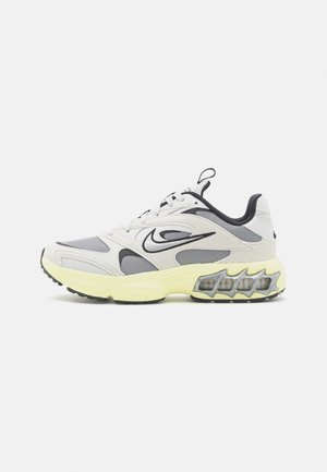 Zapatilla atlética con parte superior de malla gris y blanca, suela de goma amarilla y acentos negros. Cuenta con amortiguación visible y un patrón de tracción único.