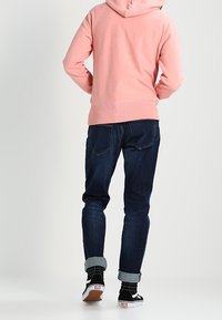 Lichtroze hoodie met capuchon en geribbelde zoom, gecombineerd met donkerblauwe jeans en zwart-witte Vans sneakers met patroon.
