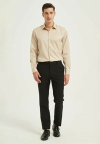 Chemise beige à manches longues avec un col pointu et une fermeture à boutons, assortie à un pantalon noir ajusté et des chaussures noires. Texture de tissu lisse.