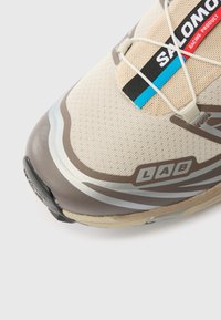 Μπεζ αθλητικό παπούτσι τρεξίματος από δίχτυ με σκουροκάστανα στοιχεία, ασημένιες λεπτομέρειες και μαύρη ετικέτα. Εξοπλισμένο με ελαστικό σύστημα κορδονιών.